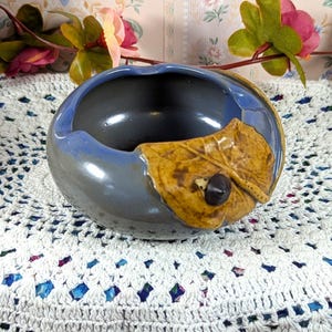 May include: A ceramic ashtray with a blue and gray exterior and a dark interior. A decorative leaf with a small black accent is on the side. The ashtray is on a white crocheted surface.