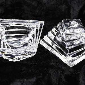 May include: Two clear, geometric glass candle holders. One is square with a circular indentation, the other is a stepped pyramid shape. Both are on a black background. The candle holders are likely for decorative use.