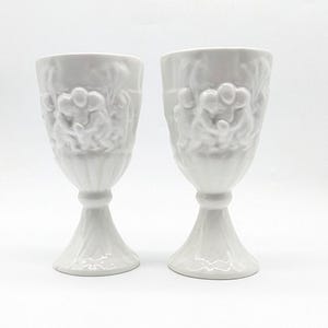 Puede incluir: Dos copas de cerámica blanca con detalles en relieve ornamentados. Las copas tienen un diseño clásico con tallo, con un cuenco ancho y un patrón de relieve detallado. Las copas son de cerámica blanca y están sobre un fondo blanco.