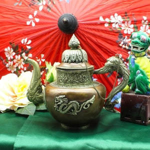 May include: A copper teapot with a dragon design and a silver lid and handle. The teapot is sitting on a green table cloth with a red and white umbrella and a green and yellow ceramic dragon figurine in the background.