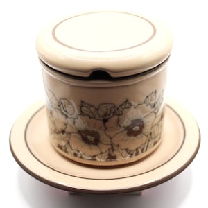 Puede incluir: Un azucarero de cerámica beige con tapa y un platillo a juego. El tazón y el platillo están decorados con un patrón floral marrón.