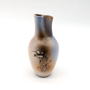 May include: A ceramic vase with a bulbous body and a narrow, slightly flared neck. The surface features a gradient of soft brown and muted blue tones, accented by a dark, etched botanical illustration of tall grasses on the front of the vessel.