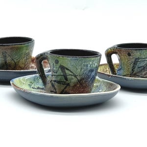 May include: Set of three ceramic espresso cups and saucers with abstract designs in shades of green, brown, and blue. The cups have black handles and the saucers are a light blue colour.