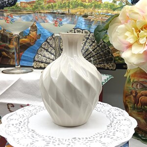 May include: A white ceramic vase with a swirling, textured design. The vase is sitting on a white doily.
