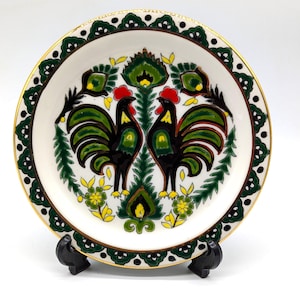 May include: A round ceramic plate with a white center featuring two black roosters with green wings and red combs facing each other. The design includes yellow flowers and green foliage. The rim has a dark green scalloped pattern with gold trim.