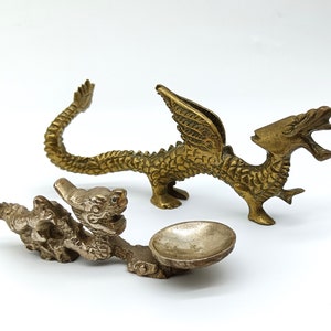 May include: Two gold-coloured dragon figurines with intricate details. One dragon has wings and a curled tail, while the other is smaller and has a bowl in its mouth.