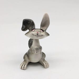May include: A ceramic figurine of a grey and white rabbit with long ears. The rabbit has a wide smile and is standing on all fours.
