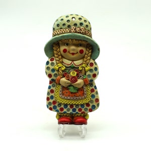 May include: A ceramic figurine of a girl wearing a blue and white polka dot hat and a colourful dress with a floral bouquet. The figurine is standing on a white stand.