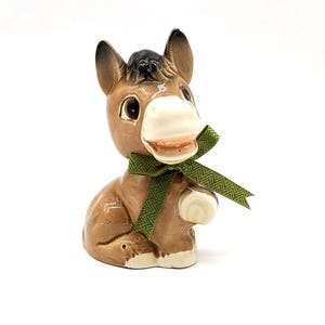 May include: A ceramic donkey figurine featuring a glossy brown finish with white accents on the muzzle, hooves, and inner ears. The donkey has large dark eyes, a black mane, and wears a green fabric ribbon tied in a bow around its neck.
