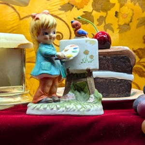 Puede incluir: Figura de cerámica de una niña con abrigo azul, sosteniendo una paleta, pintando en un pequeño caballete. Un pájaro y una cereza están encima del caballete. Una rebanada de pastel y uvas en el fondo.
