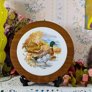 May include: A circular wall plaque featuring a white ceramic centre with a detailed illustration of a male and female mallard duck in a marshy landscape, surrounded by a wide, polished wooden frame and supported by a black plastic stand.