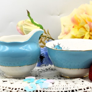 Pode incluir: Um conjunto de jarro de creme e açucareiro azul e branco com acabamento dourado. O jarro tem uma alça curva e o açucareiro é redondo. O conjunto está sobre uma toalha de renda branca com confete azul e branco.