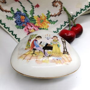 Puede incluir: Una caja de porcelana blanca con forma de concha con una escena pintada de dos personas tocando música. La escena incluye un piano, un reloj y una ventana.