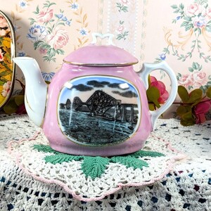 May include: A pink ceramic teapot with a lid, white spout, and handle. The teapot features a scenic image of a bridge. It sits on a white doily with a green leaf design. Decorative items are in the background.