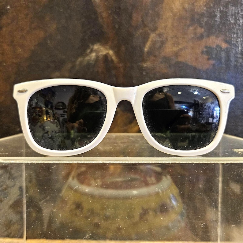Pode incluir: &Oacute;culos de sol com arma&ccedil;&atilde;o branca e lentes escuras. Os &oacute;culos de sol s&atilde;o exibidos em uma superf&iacute;cie transparente, refletindo o ambiente. O design cl&aacute;ssico apresenta uma arma&ccedil;&atilde;o grossa e uma ponte em forma de fechadura.