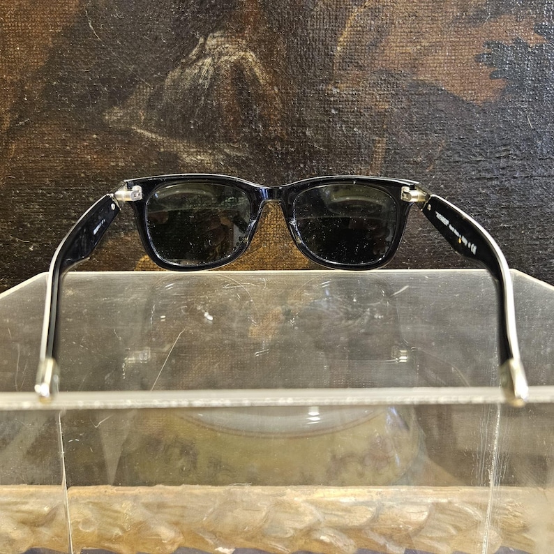 Pode incluir: &Oacute;culos de sol pretos com lentes escuras e uma arma&ccedil;&atilde;o retangular cl&aacute;ssica. As hastes s&atilde;o pretas com detalhes brancos e uma dobradi&ccedil;a prateada. Os &oacute;culos s&atilde;o exibidos em uma superf&iacute;cie transparente.