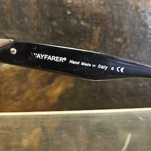 Pode incluir: Close-up de uma haste de &oacute;culos Wayfarer preta. O texto "WAYFARER&reg; Hand Made in Italy B CE" est&aacute; impresso na haste. A haste &eacute; preta com um detalhe prateado e um pequeno rebite prateado.