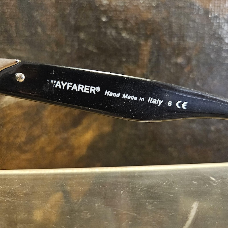 Pode incluir: Close-up de uma haste de &oacute;culos Wayfarer preta. O texto "WAYFARER&reg; Hand Made in Italy B CE" est&aacute; impresso na haste. A haste &eacute; preta com um detalhe prateado e um pequeno rebite prateado.