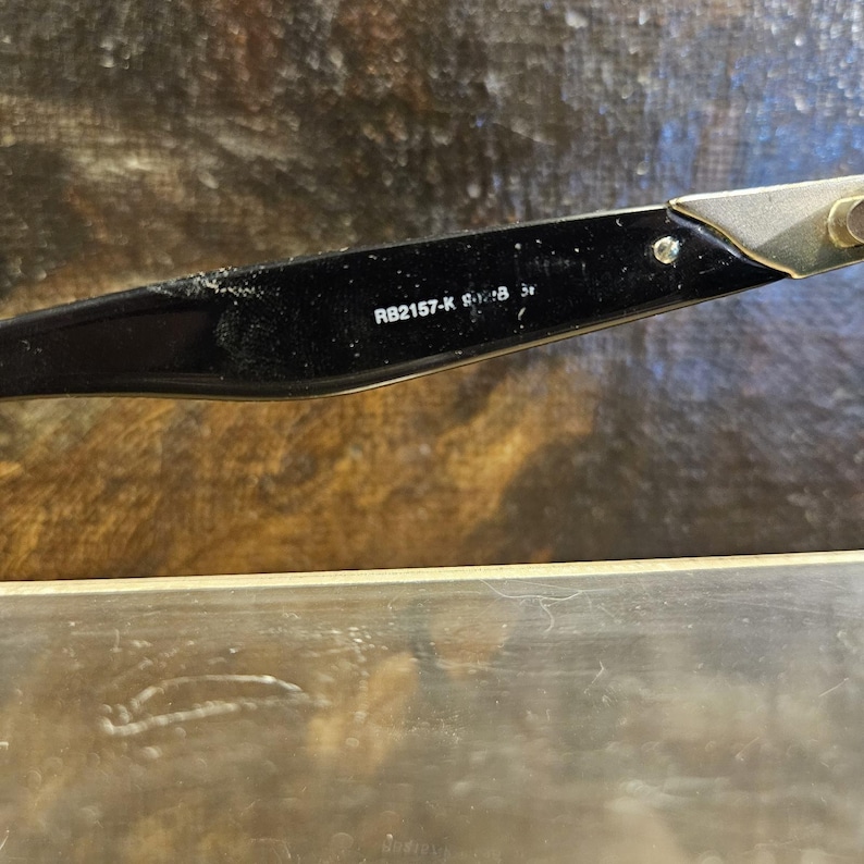 Pode incluir: Close-up de uma haste de &oacute;culos preta com o texto "RB2157-K 8-28 3r". A haste tem uma dobradi&ccedil;a de metal prateado e uma arma&ccedil;&atilde;o de pl&aacute;stico preta. O fundo &eacute; uma superf&iacute;cie desfocada e texturizada.