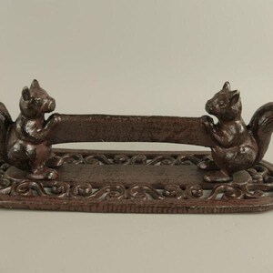May include: A cast iron squirrel-themed paper towel holder. Two detailed squirrel figures flank a horizontal bar, all mounted on an oval base with an ornate, scrollwork design. The holder has a rustic, brown finish.