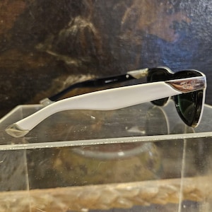Pode incluir: &Oacute;culos de sol com arma&ccedil;&atilde;o preta e hastes brancas. Os &oacute;culos t&ecirc;m um detalhe prateado e lentes escuras. O logotipo da Ray-Ban &eacute; vis&iacute;vel no detalhe prateado. Os &oacute;culos s&atilde;o exibidos em uma superf&iacute;cie transparente.
