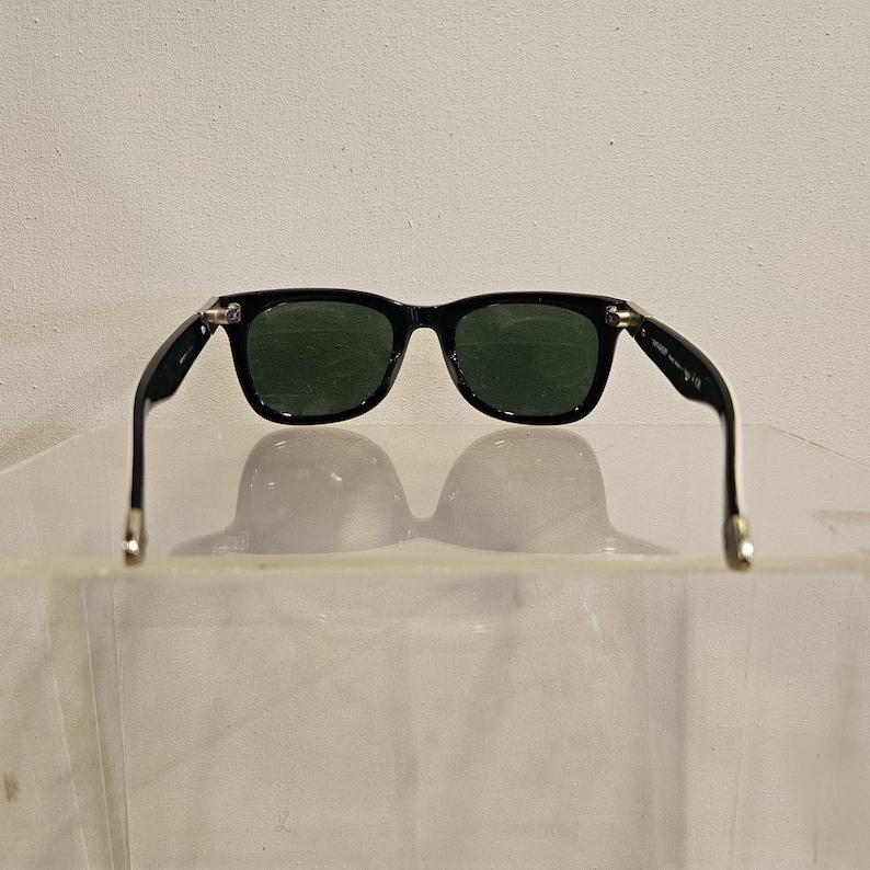 Pode incluir: &Oacute;culos de sol pretos com lentes verde escuro. Os &oacute;culos t&ecirc;m uma arma&ccedil;&atilde;o retangular cl&aacute;ssica. As hastes dos &oacute;culos s&atilde;o pretas com detalhes prateados nas dobradi&ccedil;as. Fundo branco.