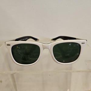 Pode incluir: &Oacute;culos de sol brancos com lentes verde escuro e hastes pretas. Os &oacute;culos de sol t&ecirc;m um design cl&aacute;ssico e retro com uma arma&ccedil;&atilde;o branca espessa. As lentes s&atilde;o de cor verde escuro e as hastes s&atilde;o pretas.