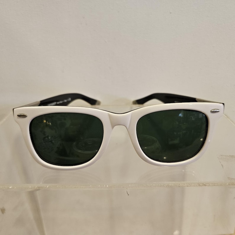 Pode incluir: &Oacute;culos de sol brancos com lentes verde escuro e hastes pretas. Os &oacute;culos de sol t&ecirc;m um design cl&aacute;ssico e retro com uma arma&ccedil;&atilde;o branca espessa. As lentes s&atilde;o de cor verde escuro e as hastes s&atilde;o pretas.