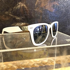 Pode incluir: &Oacute;culos de sol brancos com lentes pretas e arma&ccedil;&atilde;o branca. O logotipo da Ray-Ban &eacute; vis&iacute;vel na haste. As lentes refletem o ambiente. Os &oacute;culos de sol s&atilde;o exibidos em uma superf&iacute;cie transparente.