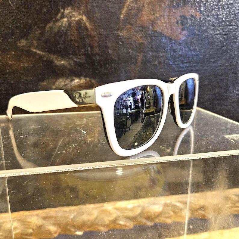 Pode incluir: &Oacute;culos de sol brancos com lentes pretas e arma&ccedil;&atilde;o branca. O logotipo da Ray-Ban &eacute; vis&iacute;vel na haste. As lentes refletem o ambiente. Os &oacute;culos de sol s&atilde;o exibidos em uma superf&iacute;cie transparente.