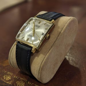 Op de afbeelding: Een vintage goudkleurig Anker automatisch polshorloge met een rechthoekige wijzerplaat en een zwarte leren band. Het horloge heeft een zilveren wijzerplaat met gouden wijzers en een datumweergave. Het horloge wordt getoond op een beige kussen.