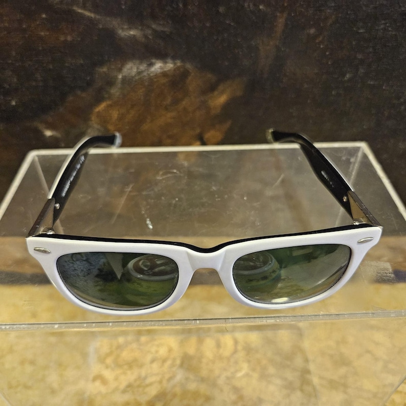 Pode incluir: &Oacute;culos de sol brancos com hastes pretas e lentes verdes. Os &oacute;culos s&atilde;o exibidos em uma superf&iacute;cie transparente. As arma&ccedil;&otilde;es s&atilde;o de estilo retr&ocirc; cl&aacute;ssico.