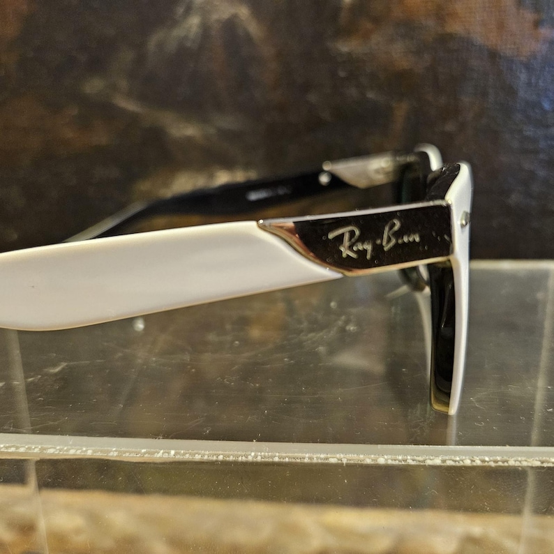 Pode incluir: Close-up de &oacute;culos Ray-Ban. As arma&ccedil;&otilde;es s&atilde;o brancas com hastes pretas e um detalhe prateado. O log&oacute;tipo Ray-Ban &eacute; vis&iacute;vel na lateral da arma&ccedil;&atilde;o. Os &oacute;culos est&atilde;o sobre uma superf&iacute;cie transparente.