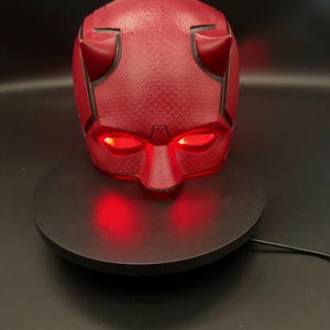 May include: A red Daredevil mask with black horns and glowing red eyes. The mask is sitting on a black circular base.