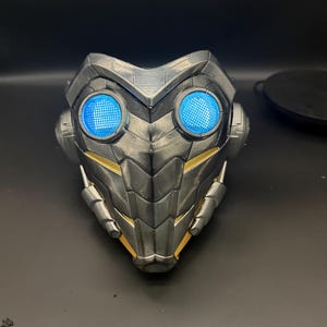 May include: A black and gold 3D printed mask with blue LED lights. The mask has a textured, scaly design and a pointed chin.
