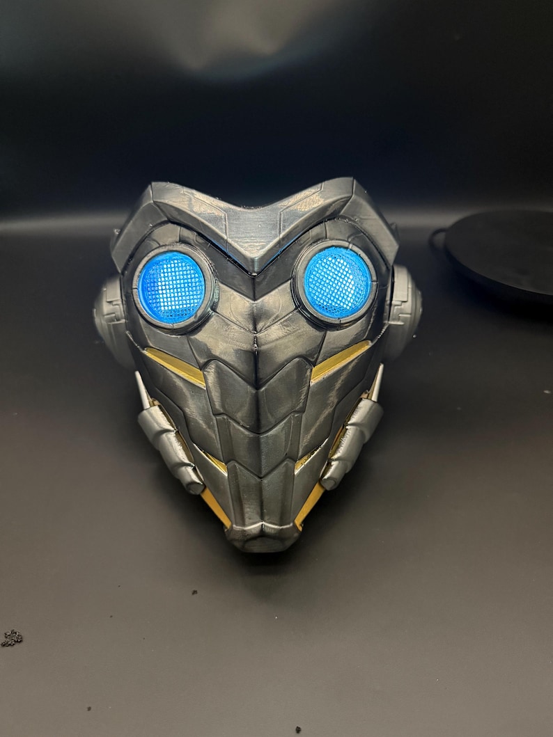 May include: A black and gold 3D printed mask with blue LED lights. The mask has a textured, scaly design and a pointed chin.