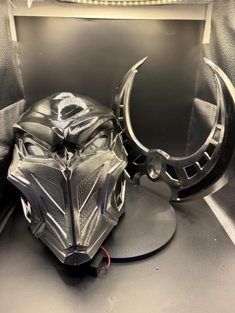 May include: A black and silver helmet with a detailed design and a matching black and silver weapon. The helmet has a visor and the weapon is a crescent-shaped blade.