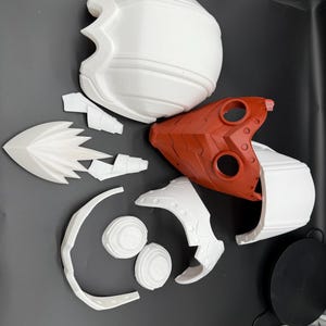 May include: A set of white and red plastic helmet pieces for cosplay or costume making. The pieces include a helmet dome, a face mask, and several smaller pieces.