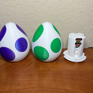 May include: Three white 3D printed egg-shaped containers with green and purple spots. The container on the right is open and holds several game cartridges.