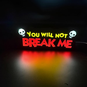 May include: Illuminated sign with the words "YOU WILL NOT BREAK ME" in red and yellow, flanked by white skull graphics. The sign glows against a dark background, with a gradient of red and orange light.