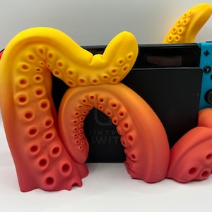 May include: A 3D printed octopus-shaped stand for a Nintendo Switch console. The stand is made of a gradient plastic that transitions from yellow to orange to red. The stand is designed to hold the console upright and has a cutout for the console's kickstand.