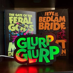 May include: A glowing neon sign with the words "GLURP GLURP" in bright green, with red and orange accents. The sign is in front of two books titled "THE GATE OF THE FERAL GODS" and "THE EYE OF THE BEDLAM BRIDE".