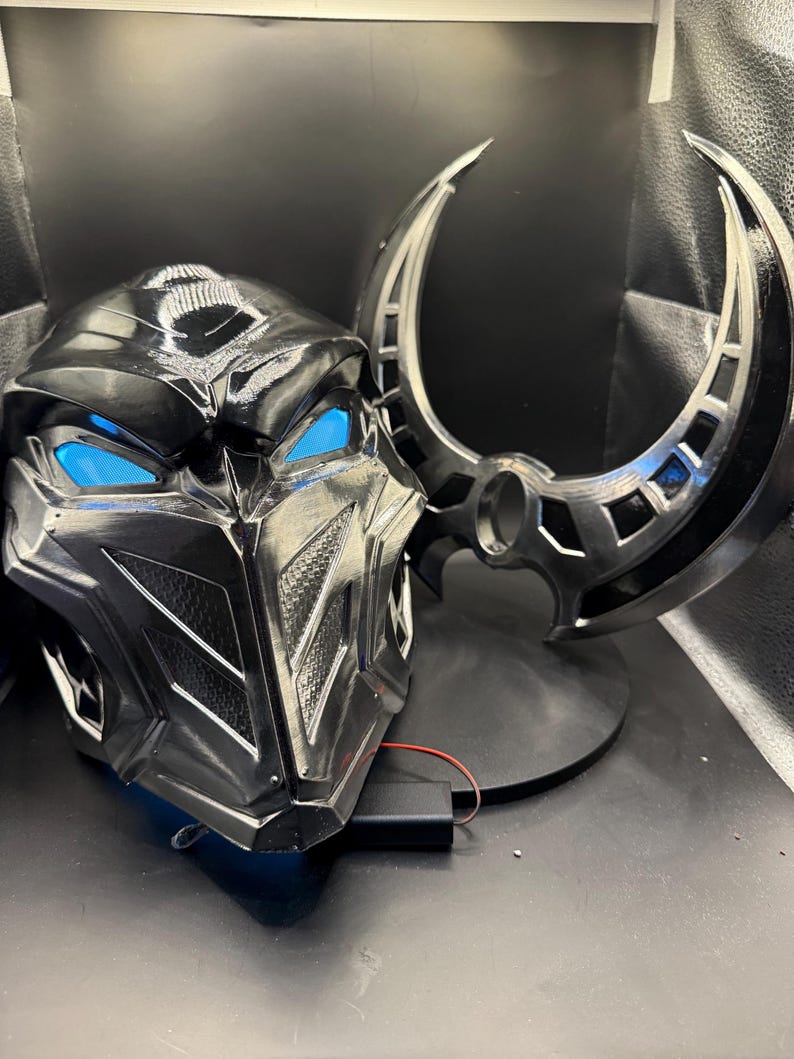 May include: A black plastic helmet with a futuristic design and blue glowing eyes. The helmet is on a black stand with a crescent-shaped object behind it.