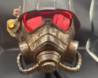 May include: A weathered, metallic tactical helmet featuring a dark gray finish with prominent rust-colored oxidation. The helmet includes glowing red eye lenses, a side-mounted yellow light, and a central respirator mask with two side hoses.
