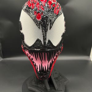 May include: A detailed mask with a menacing design, featuring a red and black color scheme. The top of the mask has a textured, red pattern, while the eyes are white with a mesh-like texture. The mouth area is red with sharp, pink teeth.