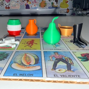 May include: A set of 3D printed game pieces including a red, white, and green barrel, an orange pitcher, a green pear, a tan bucket, a black boot, and a white skull, arranged on a colorful board game card featuring illustrations and text.