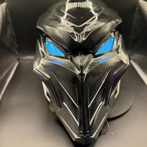 May include: A black plastic mask with a futuristic design. The mask has a sharp, angular shape with a prominent beak and two blue glowing eyes.