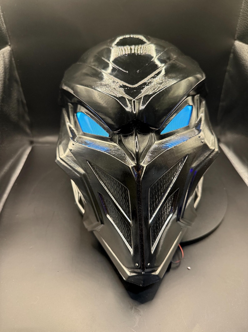 May include: A black plastic mask with a futuristic design. The mask has a sharp, angular shape with a prominent beak and two blue glowing eyes.