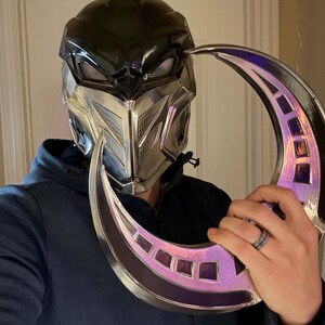 May include: A silver and black helmet with a crescent-shaped blade. The helmet has a black visor and a silver faceplate with a geometric design. The blade is silver with a purple iridescent finish.