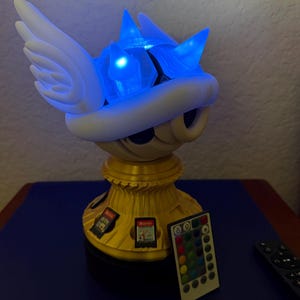 May include: A white and gold light-up figurine of a winged Koopa shell from the Super Mario Bros. video game series. The figurine has a blue light that shines from the shell and a remote control for changing the light color. The base of the figurine has slots for storing Nintendo Switch game cartridges.
